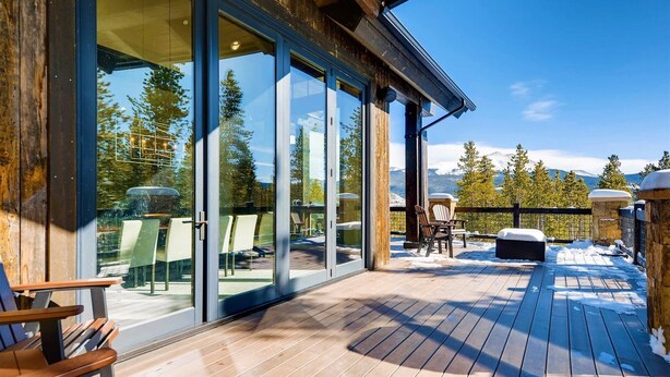 Una terraza de madera con puertas de cristal que dan a un área de descanso, una chimenea de piedra y vistas de montañas nevadas.