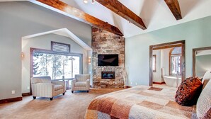 A spacious bedroom with a large bed, a stone fireplace, a television, and a view of the outdoors. - Breck_grand_vista_lodge (Breckenridge)