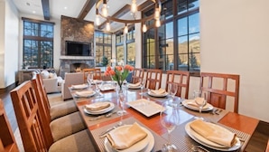 A dining area with a long wooden table set for a meal, flanked by wooden chairs. A fireplace is visible in the background, and the room has large windows offering a view of the outdoors. - Vail_alpenzauber_lodge_5bd (Vail)