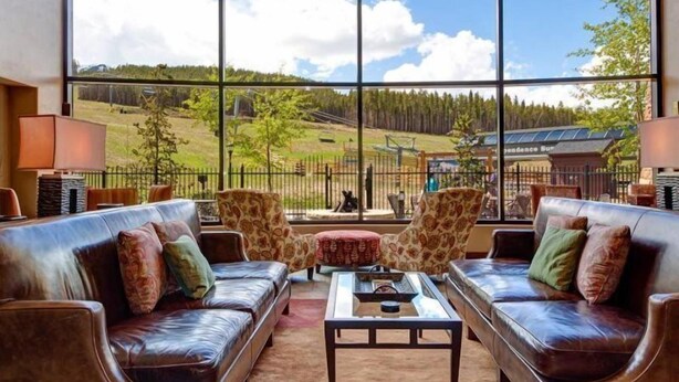 A spacious living area with leather sofas, a coffee table, and a large window offering a view of a forested landscape and a building labeled "Independence Sun." - Crystal Peak 2 Bedroom Condo at Peak 7 (Breckenridge)