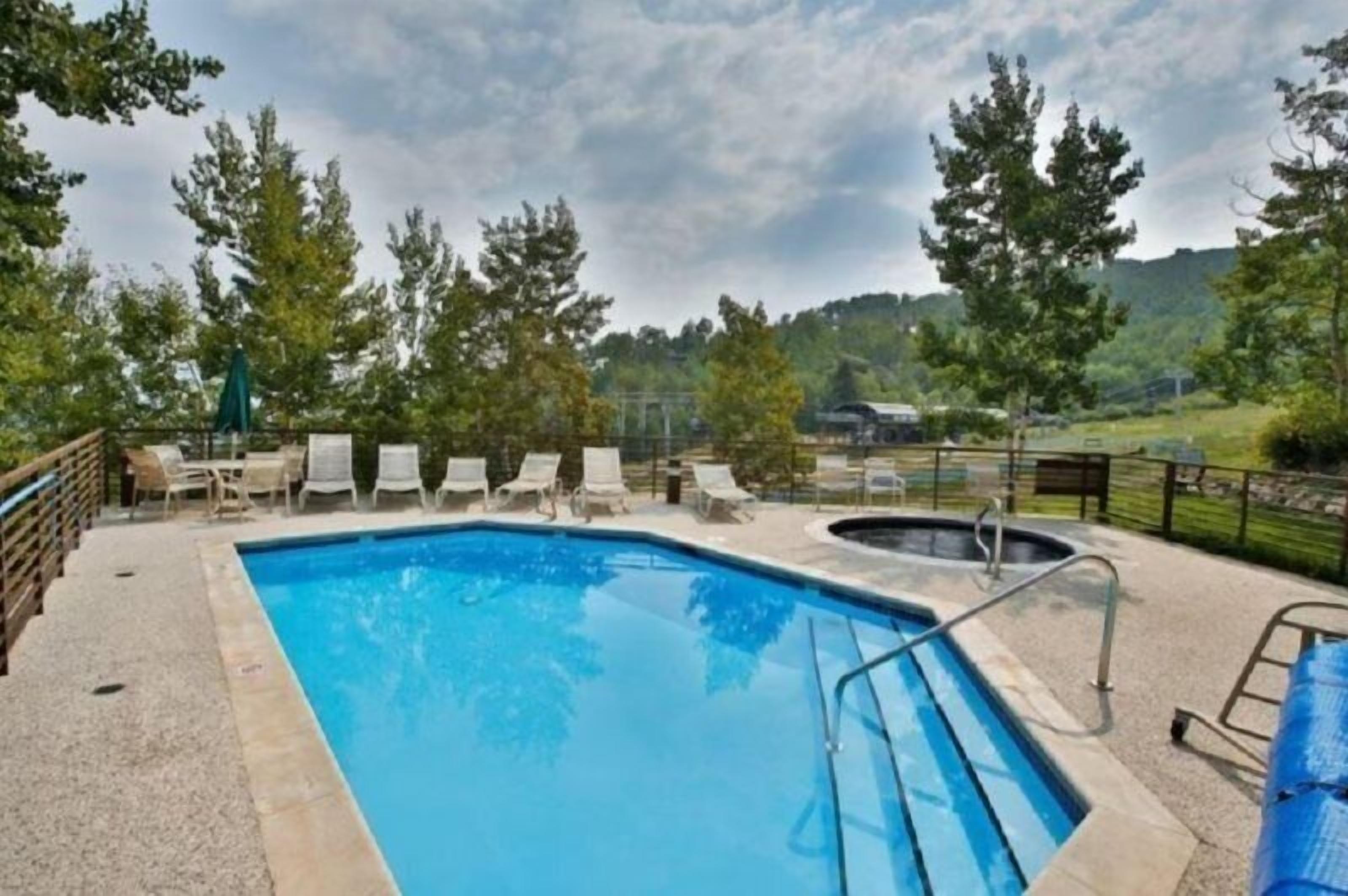 A swimming pool with lounge chairs and a hot tub, surrounded by trees and a view of hills.