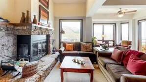 A living room with a stone fireplace, a sofa set, a coffee table, and large windows offering a view. - Snowmass_village_3bd_gen_c (Snowmass Village)