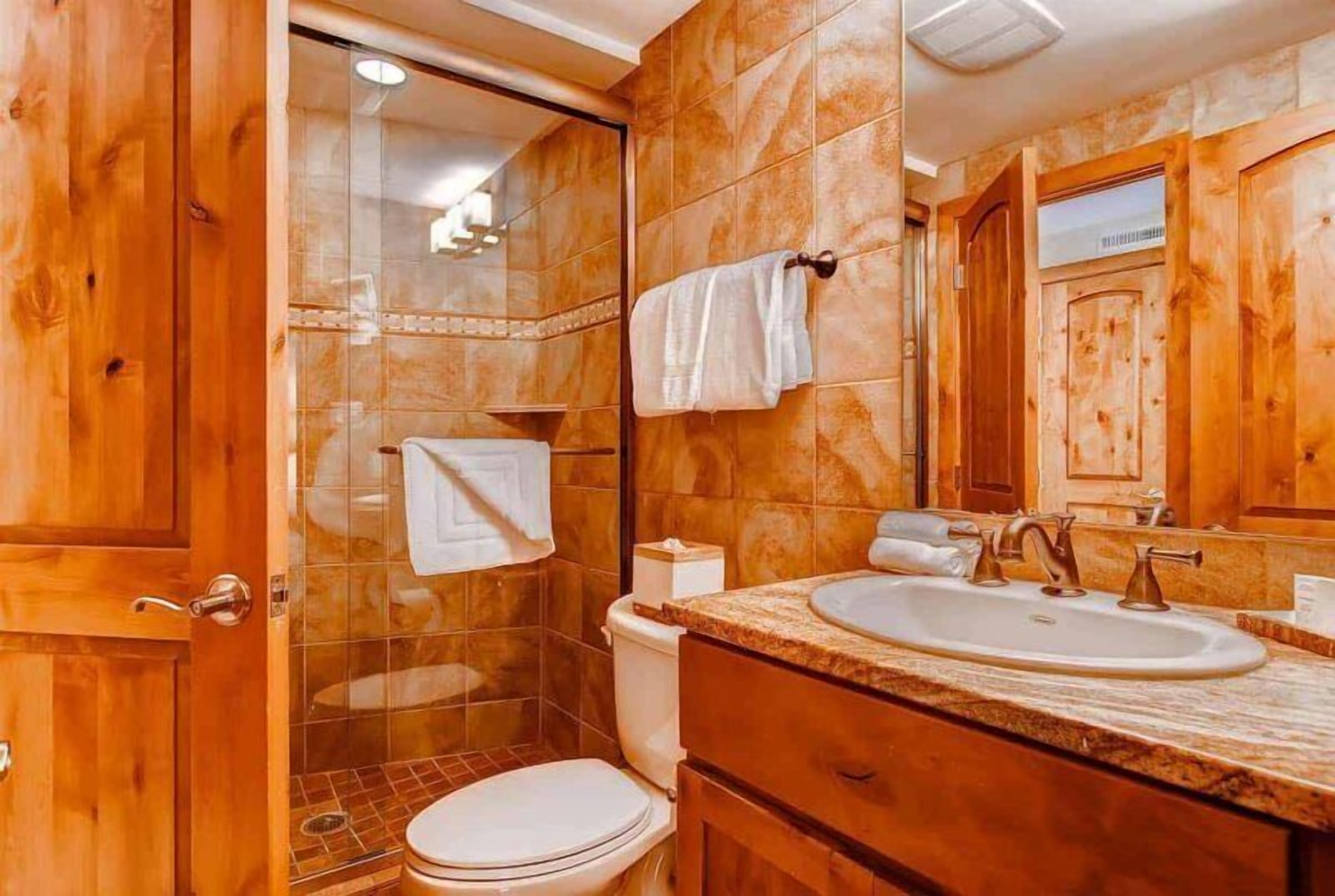 A bathroom with wooden cabinets, a glass shower enclosure, a white toilet, and a sink with a marble countertop.