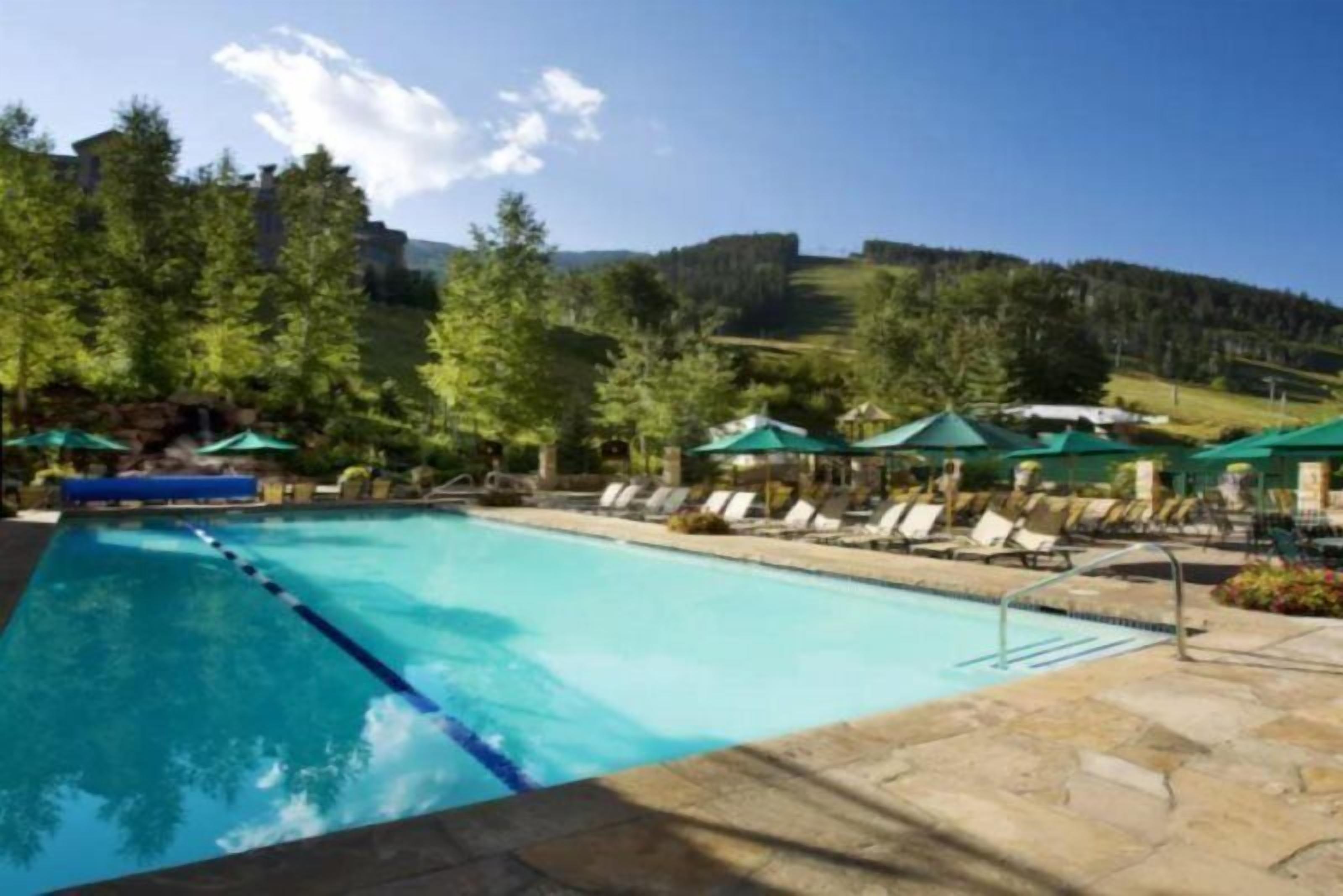 Piscina con sillas de descanso y sombrillas, rodeada de árboles y un paisaje montañoso.