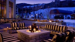 A cozy outdoor seating area with striped sofas and chairs, a stone fire pit, and a view of a snowy mountain resort in the background. - Beaver Creek Park Hyatt Double Queen Hotel Room (Avon)