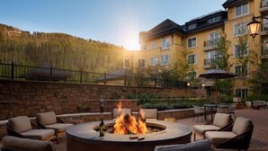 A circular outdoor seating area with a fire pit, surrounded by wicker chairs and tables, set against a backdrop of a hotel building and a forested hillside. - Vail Ritz-carlton 2 Bedroom Residence (Vail)