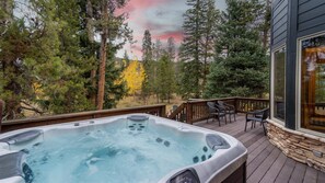 Ein Whirlpool auf einer Holzterrasse mit Stühlen, umgeben von Bäumen und einem Haus mit großen Fenstern.