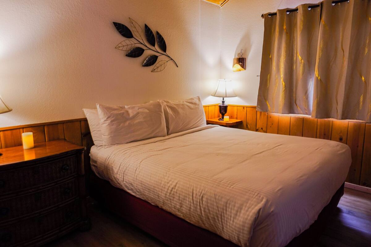 Ein Schlafzimmer mit einem hölzernen Kopfende, einem Bett mit weißer Bettwäsche, einem Nachttisch mit Lampe und einer wandmontierten Blattschmuck.