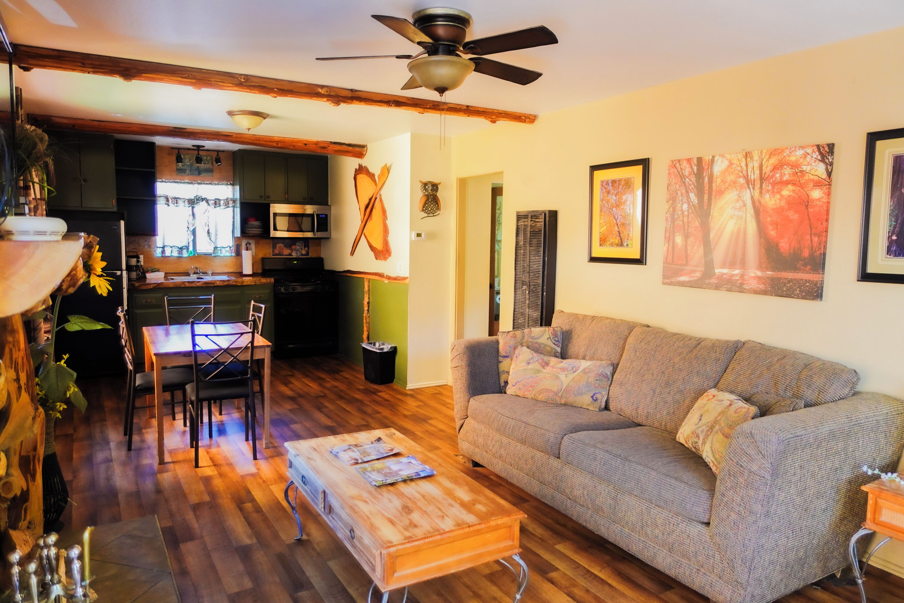 A cozy living room with a sofa, coffee table, and dining area. There is a kitchen in the background with a microwave and cabinets.