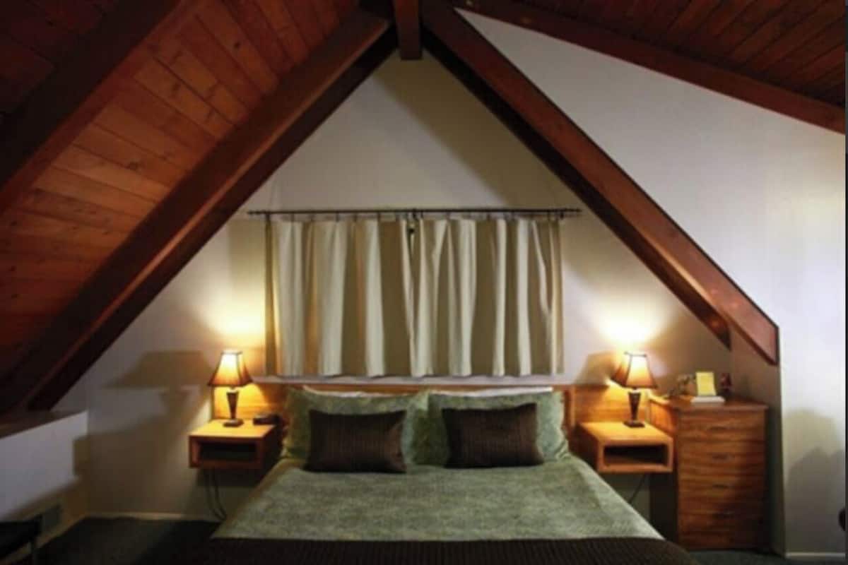 Ein Schlafzimmer mit schrägverdeckter Decke, einem Bett mit grüner Decke, zwei Nachttischen mit Lampen und einem Fenster mit Vorhängen.