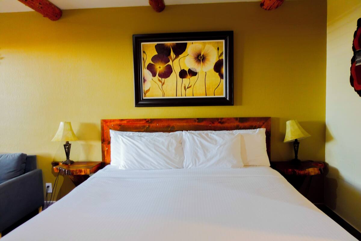 Ein Hotelzimmer mit einem Bett, zwei Nachttischen mit Lampen, einem gerahmten Blumenbild an der Wand und einem dunkel gefärbten Sessel.