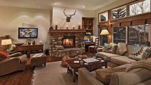 A cozy living room with a stone fireplace, comfortable seating, a television, and a large window with a view of trees. - Beaver_creek_chalet_verve (Avon)