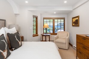Quarto com cama, poltrona, luminária, cômoda e janela com vista.