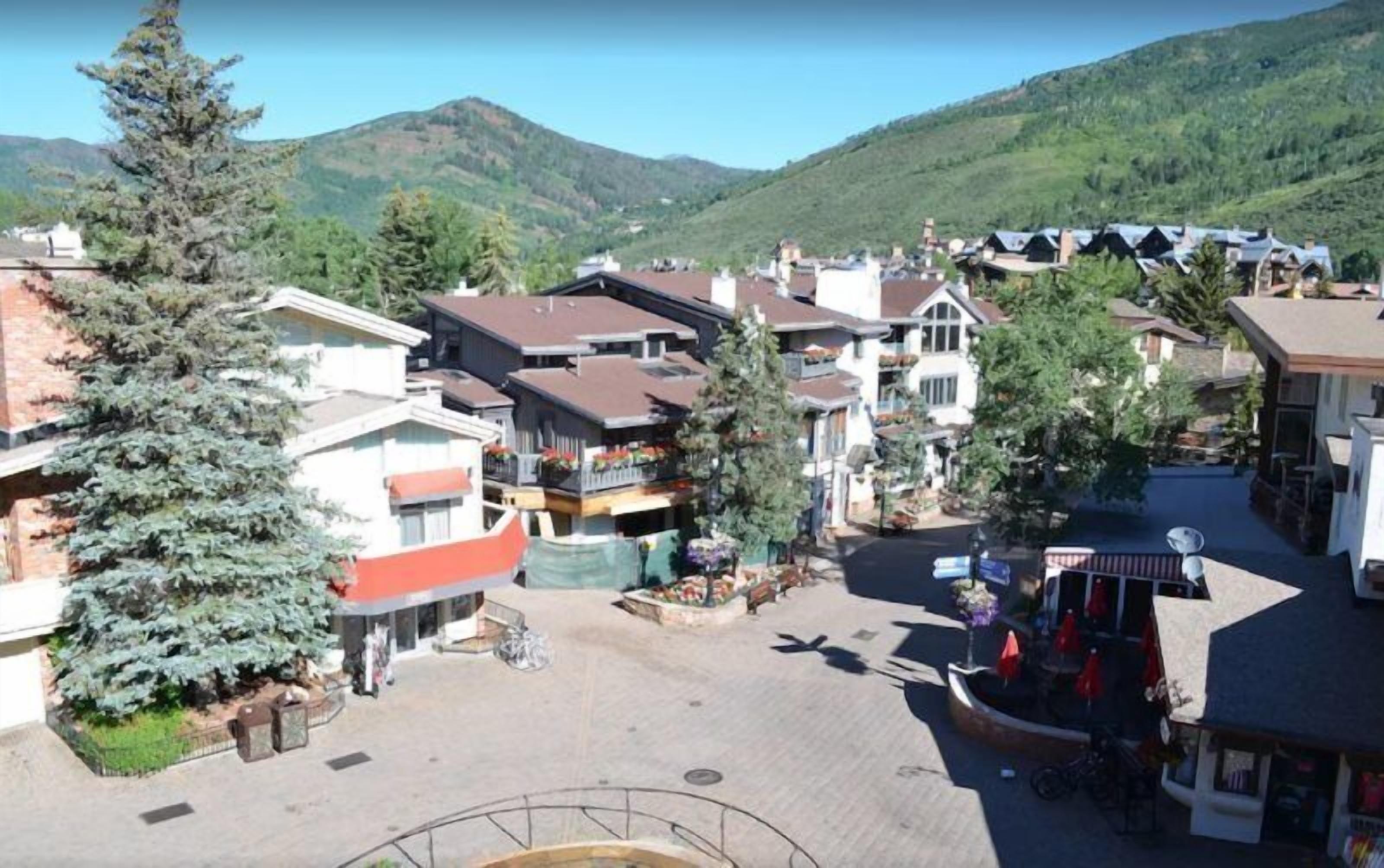 Una plaza de pueblo con edificios, un árbol grande y una montaña al fondo.
