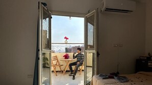 Ένα δωμάτιο με ένα κρεβάτι, μια συρόμενη γυάλινη πόρτα που οδηγεί σε μια εξωτερική βεράντα με ένα τραπέζι και καρέκλες, και μια μονάδα κλιματισμού στον τοίχο.