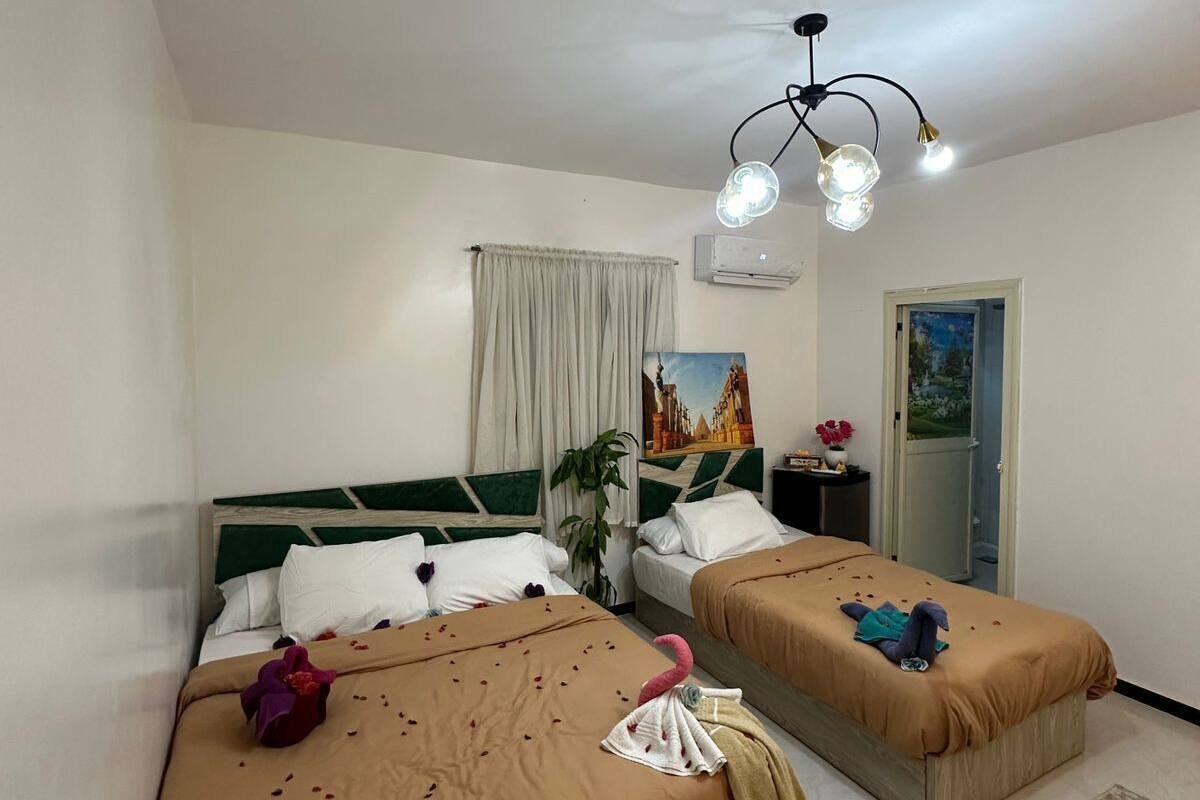 Ein Hotelzimmer mit zwei Betten, einem Deckenventilator, einer Klimaanlage, einem Fenster mit Vorhängen, einem Gemälde und einer Tür zu einem Balkon.