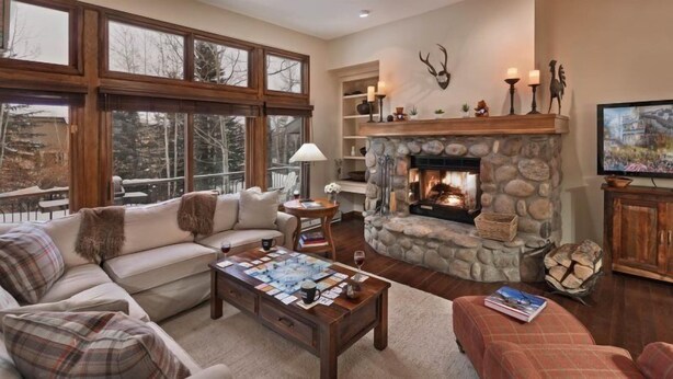 A cozy living room with a stone fireplace, a sofa, a coffee table with magazines, and a television on a wooden cabinet. - Beaver_creek_chalet_fortessa (Avon)