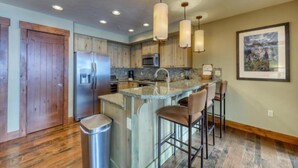 A kitchen with wooden cabinets, a stainless steel refrigerator, a granite countertop, and a bar area with stools. - One Ski Hill Place 2 Bedroom Luxury Condo (Breckenridge)