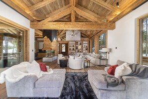 A spacious living area with high ceilings and exposed wooden beams, featuring a fireplace, comfortable seating, and large windows. - Breck_le_mayen _6bd (Breckenridge)