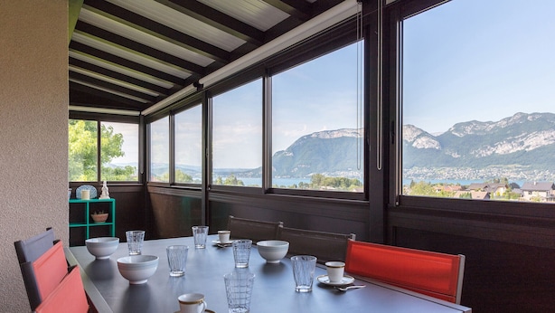 A dining area with a table set for four, large windows with a mountain view, and a green shelf with decorative items. - SEVRIER, Maison , VUE Panoramique sur le lac et montagnes, 8pers (Sévrier)