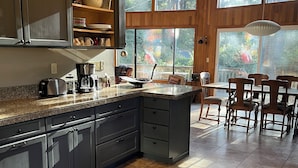 Una cocina moderna con armarios oscuros, encimera de granito y una isla grande. Hay una zona de comedor con sillas de madera y vistas a árboles a través de ventanas amplias.