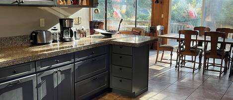 A modern kitchen with dark cabinets, a granite countertop, and a large island. There is a dining area with wooden chairs and a view of trees through large windows.