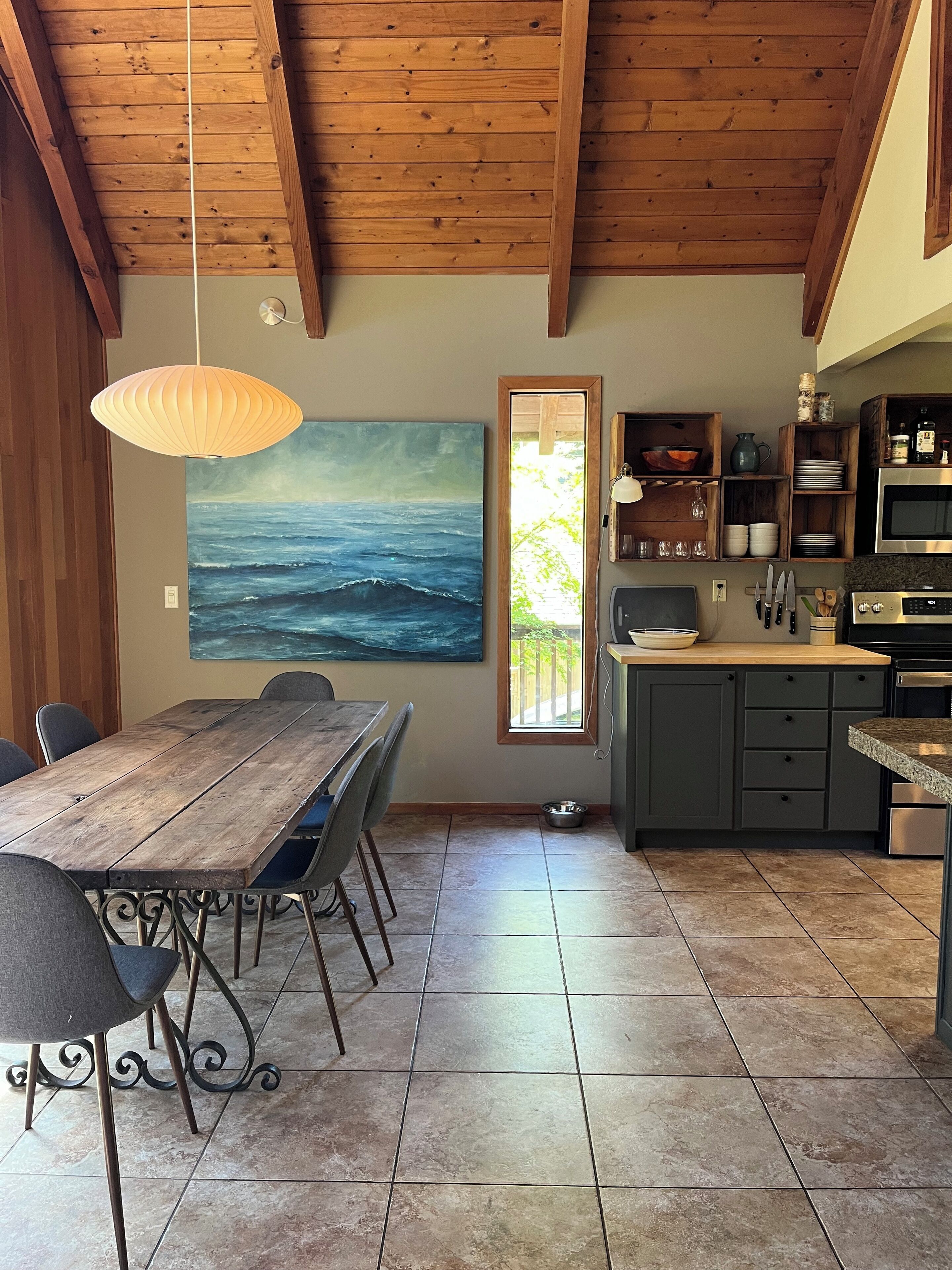 Uma sala de jantar com mesa e cadeiras de madeira, um quadro do oceano na parede e uma cozinha com eletrodomésticos modernos.