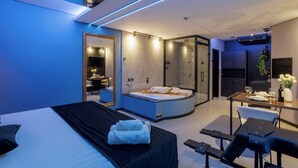 A modern hotel room with a large bed, a separate bathroom with a jacuzzi, a dining area with a table and chairs, and a TV mounted on the wall. - Medieval Motel SPA (Porto Alegre)