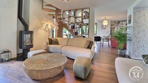 A modern living room with a sofa, ottoman, and round coffee table. There is a fireplace, bookshelves, and a dining area in the background. - My Cottage in Canada (GOSNE)