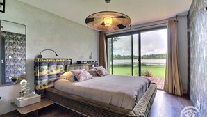 A bedroom with a large bed, a bedside table, a mirror, and a view of a lake through large windows. - My Cottage in Canada (GOSNE)