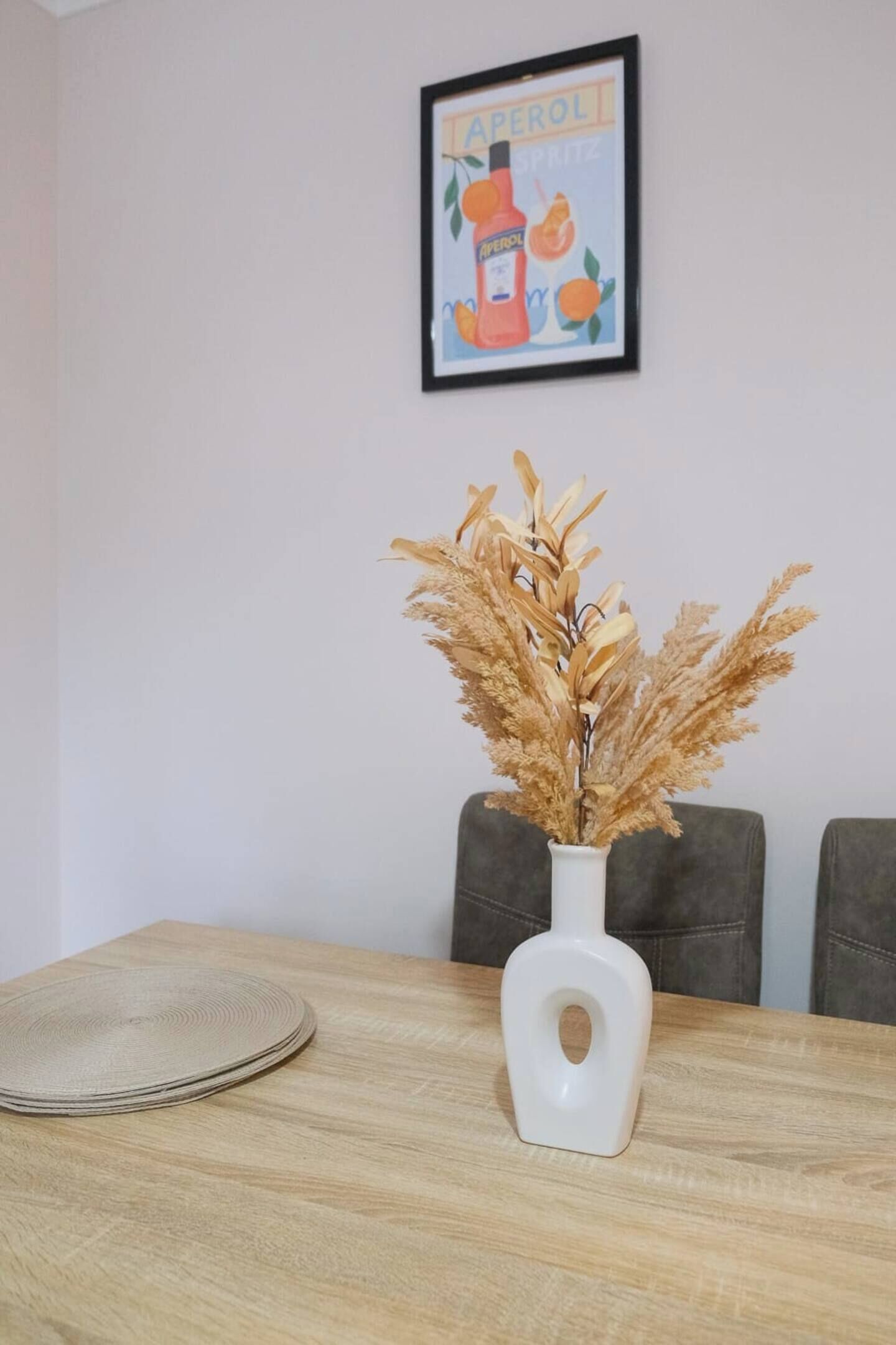 Een eettafel met een witte vaas met gedroogde planten, een ingelijste poster aan de muur, en een paar grijze stoelen.