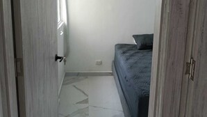 Ein schmaler Flur führt zu einem Schlafzimmer mit Bett, Fenster und einer Klimaanlage an der Wand.
