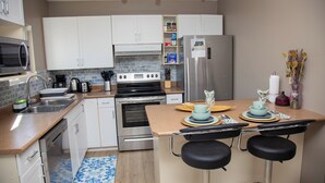 Une cuisine moderne avec des armoires blanches, des appareils en acier inoxydable, un four à micro-ondes, une cuisinière et un réfrigérateur. On y trouve deux tabourets de bar et un petit îlot central doté d’un évier et d’une bouilloire.