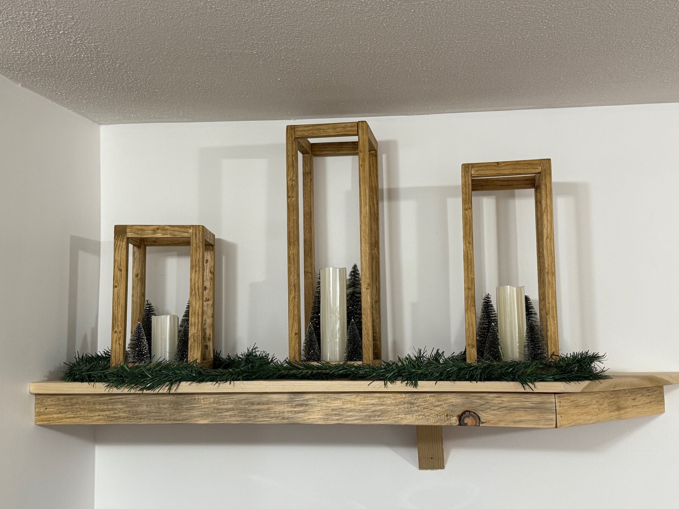 Un estante de madera con tres marcos rectangulares que contienen pequeños árboles de Navidad y velas.