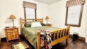 Une chambre à coucher avec un lit en bois, deux tables de chevet, un ventilateur de plafond et une fenêtre agrémentée d’une lambrequin à motifs floraux.