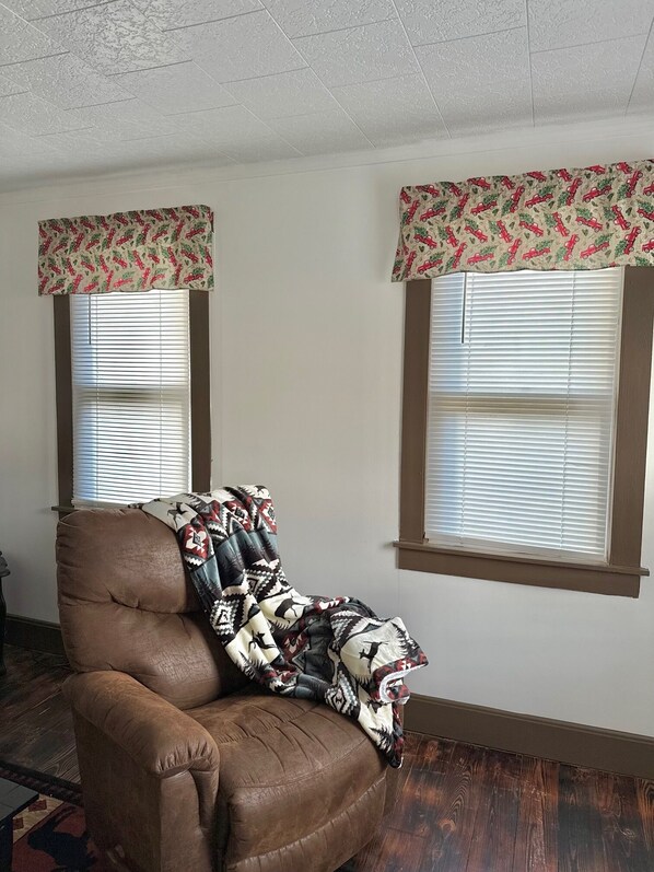 A brown leather armchair with a patterned throw blanket, positioned in front of a window with blinds and a floral valance.