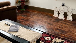Une pièce dotée d’un ventilateur de plafond, de deux fenêtres ornées de rideaux aux motifs festifs, d’une échelle en bois, d’une petite étagère décorative et d’un téléviseur fixé au mur.