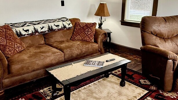 Un salon avec un canapé en cuir marron, un fauteuil assorti, une table basse, un tapis à motifs et une lampe fixée au mur.