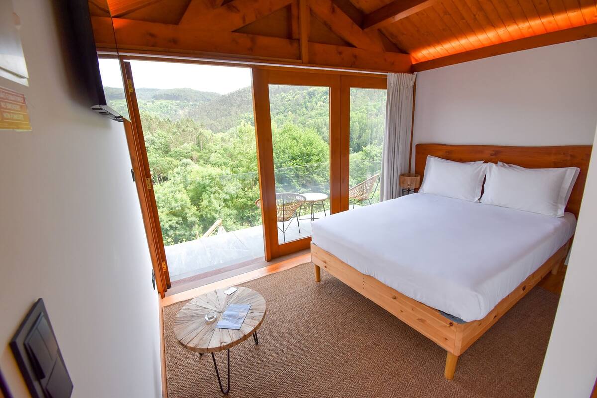 Een slaapkamer met een houten bed, een klein rond tafeltje en uitzicht op een bebost landschap door een groot raam.