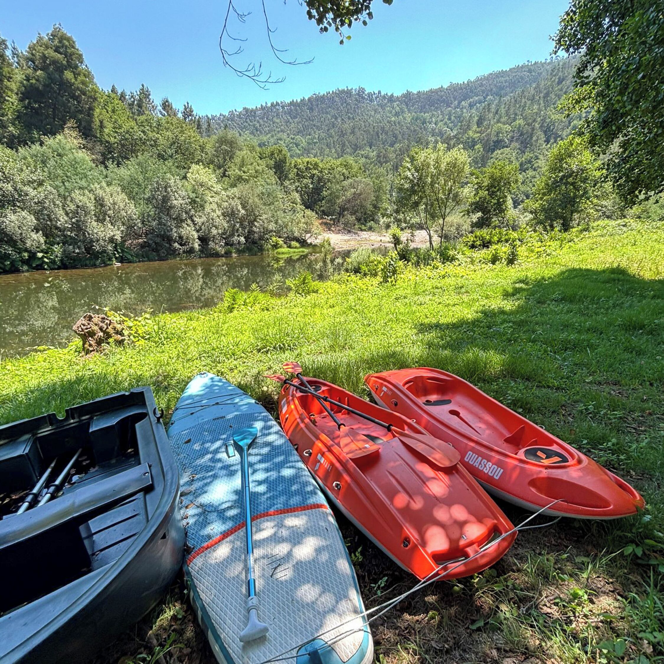 Kayaks and a canoe by a river with a forested hillside in the background.