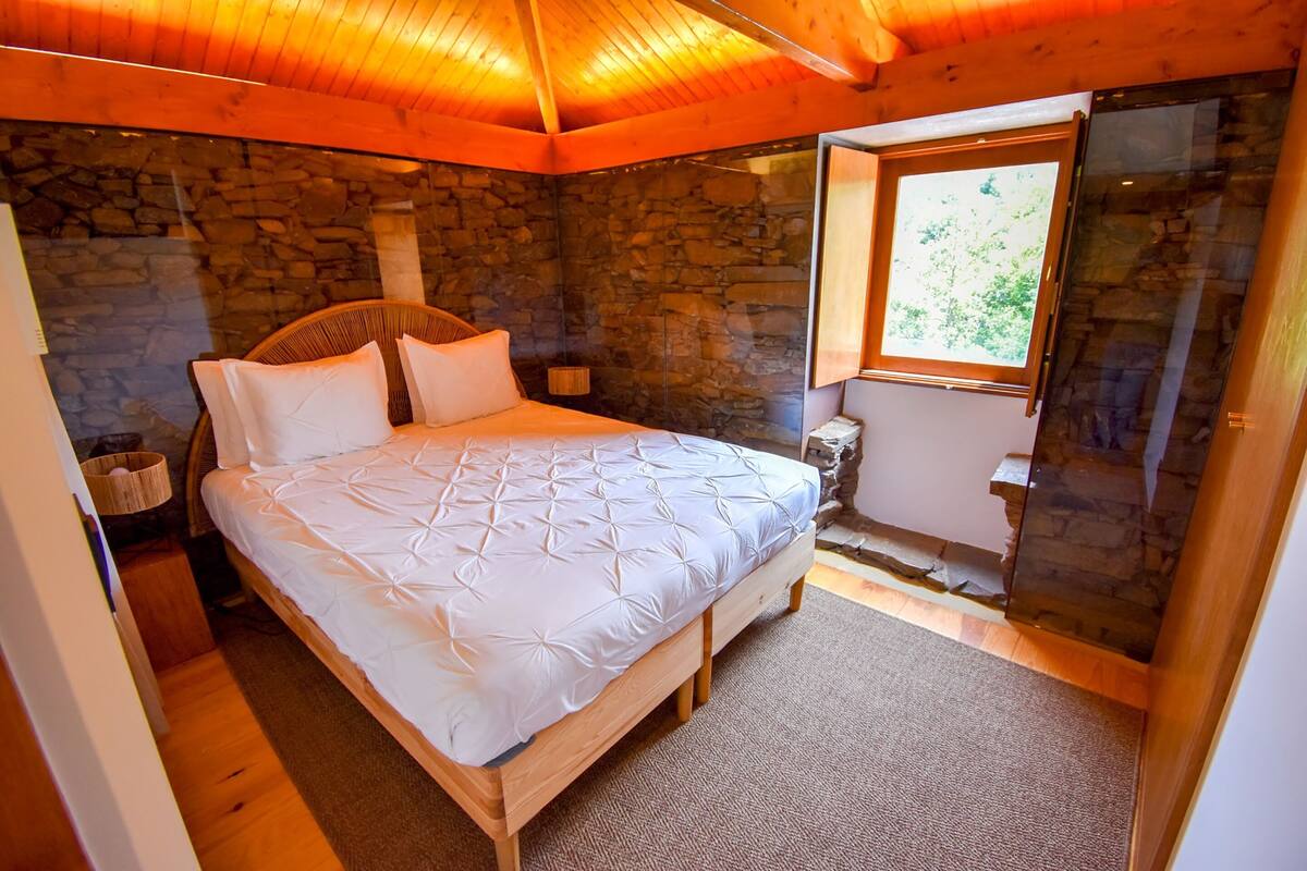 Een slaapkamer met een stenen muur, een houten plafond, een bed met witte beddengoed, een rieten stoel en een klein raam.