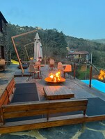 Un patio con bancos y mesas de madera, un brasero y vistas a una piscina y colinas.