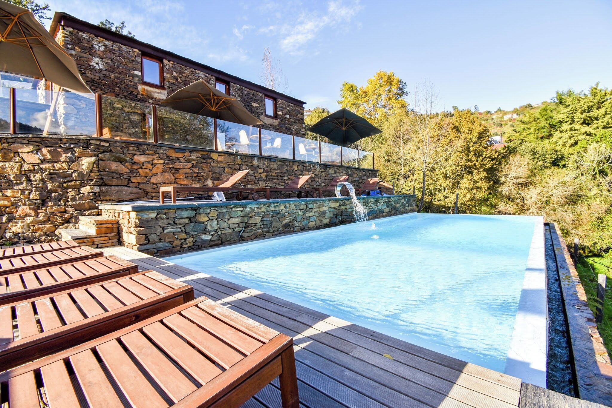 Un edificio de piedra con piscina, terraza de madera y sillas de descanso.