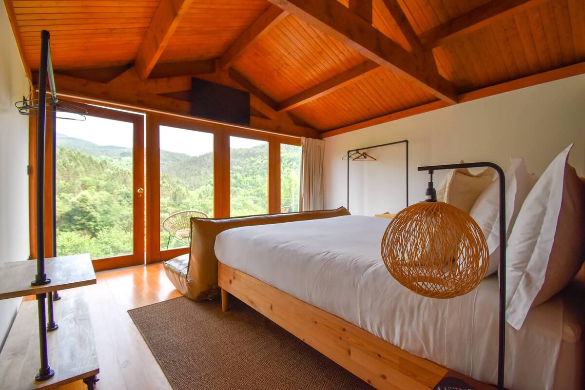 Een slaapkamer met een groot bed, houten balken in het plafond, uitzicht op het bos en een tafel met een lamp.