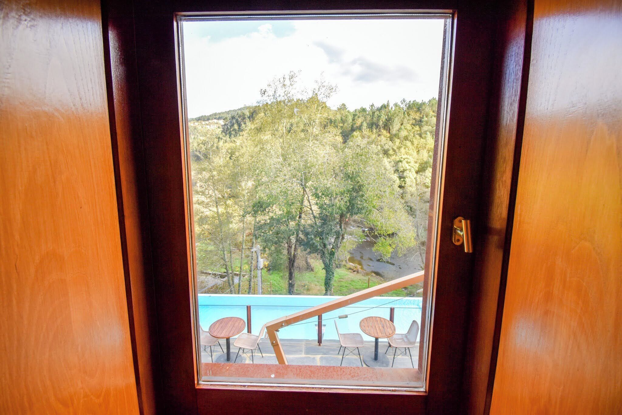 A wooden door with a view of a pool area, outdoor seating, and a forested hillside.