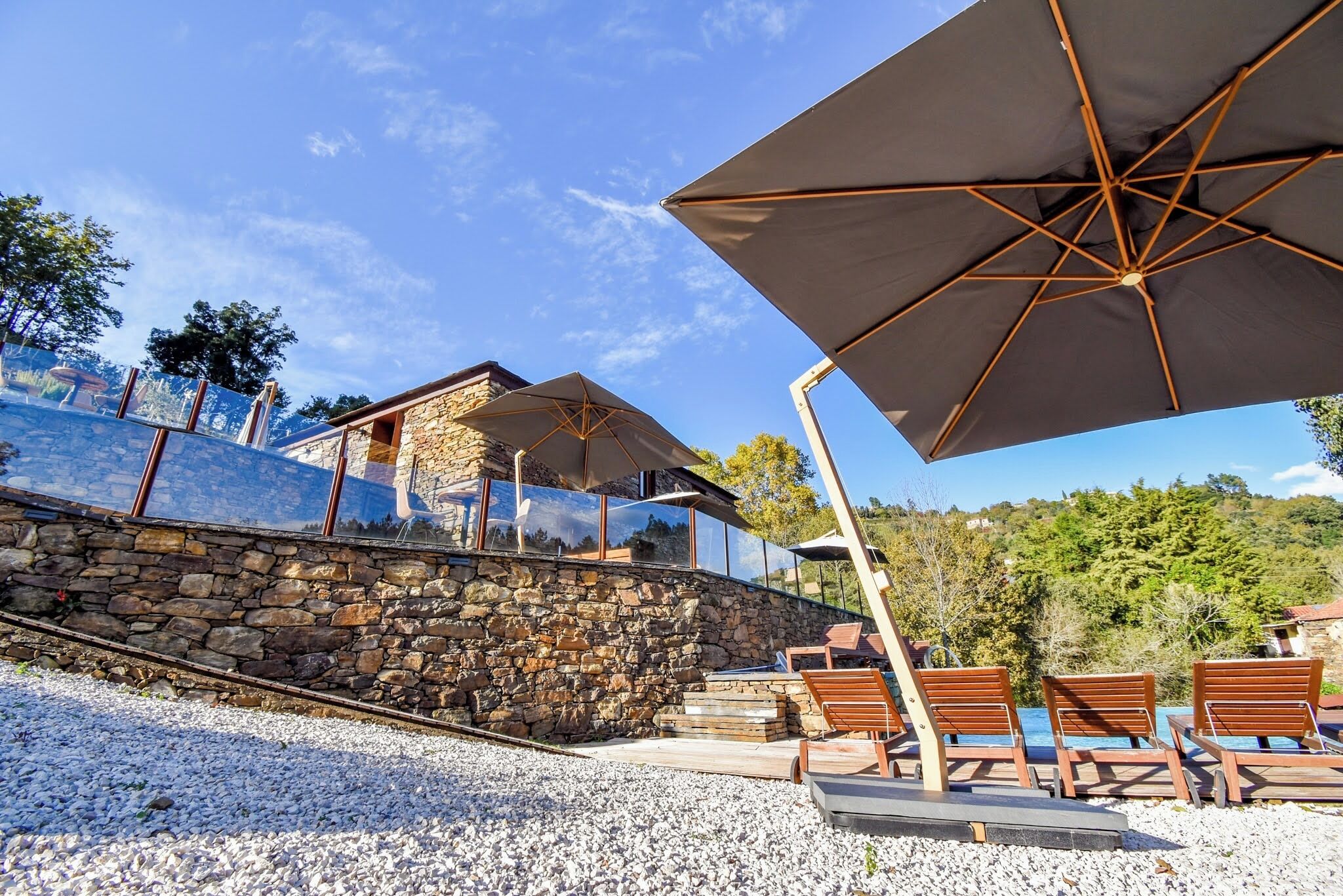 A stone wall with a glass balustrade, outdoor seating with umbrellas, and a view of a mountain.