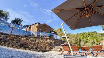 A stone wall with a glass balustrade, outdoor seating with umbrellas, and a view of a mountain.