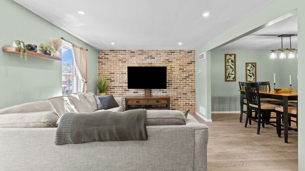 A living room with a brick wall, a flat-screen TV, a sofa, a dining table with chairs, and a hanging light fixture. - Yinzer On The Mount - 5 Min. To Downtown  (Pittsburgh)