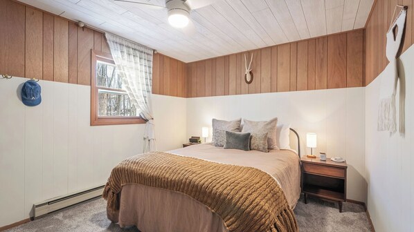 A bedroom with a bed, bedside tables, a lamp, a heater, a window with curtains, and a wall clock. - The Pines Cozy Arrowhead Lake Escape With Pools and Outdoor Adventures (Pocono Lake)