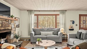 A cozy living room with a stone fireplace, a grey sofa, a round coffee table, and a view of trees through large windows. - The Pines Cozy Arrowhead Lake Escape With Pools and Outdoor Adventures (Pocono Lake)